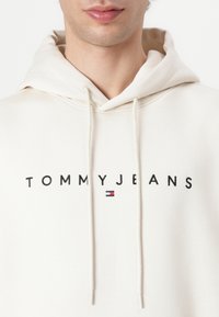 Crème-kleurige hoodie met een trekkoord, voorzien van het "TOMMYJEANS" logo in het zwart en een klein rood, wit en blauw embleem. Gemaakt van een katoenmix.