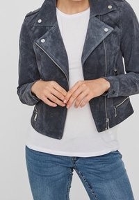 Grå mockajacka i motorcykelstil med silverdetaljer, dragkedjefickor och skärskjortslag, tillsammans med en vit t-shirt och blå jeans.