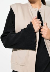 Beżowa kamizelka bez rękawów z zapięciem na zatrzaski, w połączeniu z czarnym prążkowanym topem z długim rękawem, ozdobionym rozszerzanymi mankietami i wieloma pierścieniami.