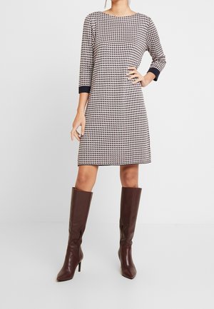 Femme portant une robe noir et blanc à motif pied-de-poule, longueur genou, manches trois-quarts, et des bottes marron montantes à talons.