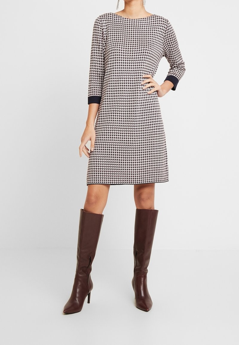 Femme portant une robe noir et blanc à motif pied-de-poule, longueur genou, manches trois-quarts, et des bottes marron montantes à talons.