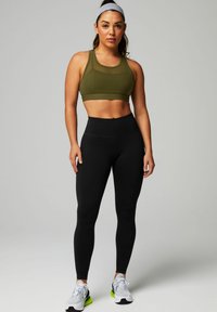 Olivgrüner Sport-BH kombiniert mit hoch-taillierten schwarzen Leggings. Das Model trägt weiße und grüne Sportschuhe. Glatter Stoff mit Stretch.