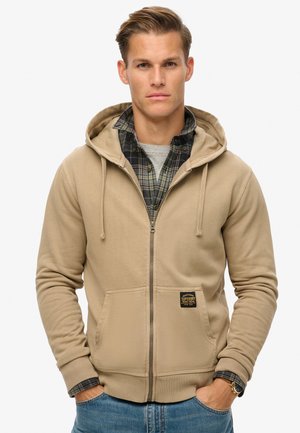 Beige zip-up hoodie met een kangoeroezak, trekkoorden, geribbelde manchetten en een logo-patch. Gemaakt over een geruite shirt en een grijs T-shirt.