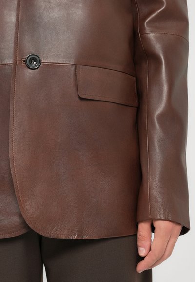 Brown leather jacket with buttoned flap pocket and black button, worn over dark pants, with a hand relaxed at the side.