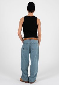 Homme portant un bonnet noir, un débardeur noir, un jean bleu clair à jambes larges, une ceinture marron et des chaussures marron, debout de dos sur un fond uni.