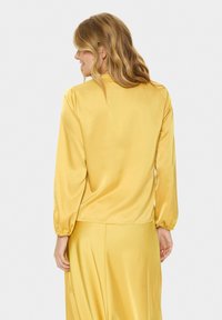 Gelbe Satinbluse mit langen Puffärmeln, glatter Textur und rundem Ausschnitt. Der Rücken zeigt ein leicht gerafftes Detail am Saum.