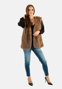 Gilet in finta pelliccia marrone con design senza maniche. Abbinato a un top nero e jeans skinny blu. Tacchi alti neri completano l'outfit.