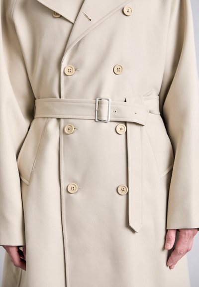 Trench coat beige doppiopetto con grandi bottoni, vita con cintura e tasche laterali. Realizzato in un tessuto liscio e texture.