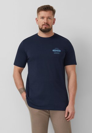 Mężczyzna z brodą w granatowej koszulce z krótkim rękawem z małym niebieskim logo na klatce piersiowej i beżowych spodniach, stojący na szarym tle.