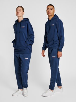 Deux mannequins portant des sweats à capuche bleu marine assortis et des pantalons de survêtement avec des baskets blanches, debout devant un fond clair uni.