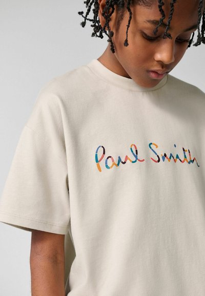 Ung person med kort krøllet hår iført en beige T-shirt med et farverigt, skråskrift "Paul Smith"-logo på brystet.
