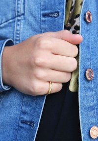 Gouden ring met een gestructureerde band en kleine ingelegde stenen, gedragen op een hand tegen een denimjack met koperen knopen en een zwart shirt.