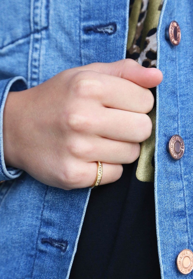 Gouden ring met een gestructureerde band en kleine ingelegde stenen, gedragen op een hand tegen een denimjack met koperen knopen en een zwart shirt.