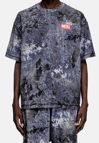 Muž v odpovídajícím modro-černém žíhaném tričku s krátkým rukávem a šortkách, s červenou nášivkou loga Diesel na hrudi a na šortkách.