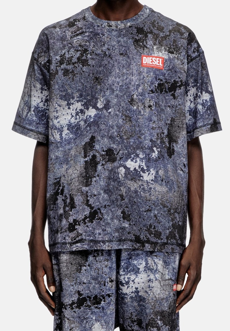 Muž v odpovídajícím modro-černém žíhaném tričku s krátkým rukávem a šortkách, s červenou nášivkou loga Diesel na hrudi a na šortkách.