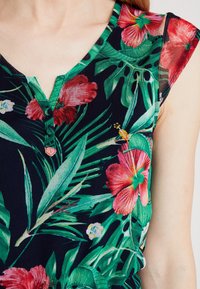 Yosamiskorinen toppi tummalla taustalla, jossa on punaisia hibiskuksia ja vihreitä lehtiä. V-aukko, jossa on kolme nappia ja lyhyet hihat.