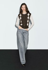 Woman with long curly hair wearing a brown argyle sleeveless cardigan over a white long-sleeve top, light blue wide-leg jeans, and black shoes.