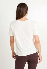 Camiseta de algodón color crema con mangas enrolladas, ajuste relajado y escote redondeado. La tela parece suave con una textura lisa.