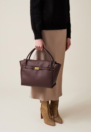 Borsa in pelle marrone con finitura testurizzata, dettagli in oro e due manici corti, tenuta contro uno sfondo di un outfit neutro.