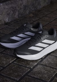 Baskets de course Adidas en noir et blanc posées sur un sol en pavés texturés, montrant les vues latérale et supérieure des deux chaussures.