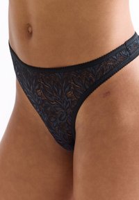 Schwarzer Spitzen-Tanga mit Blumenmuster, mit gewellten Kanten und einem dünnen Bund. Ein Etikett an der Seite zeigt die Nummer 225.