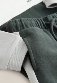I pantaloni jogger verde scuro presentano un tessuto texturizzato con un elastico in vita. I polsini sono in cotone ribbing grigio chiaro, creando un contrasto con il colore principale.