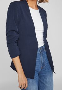Blazer bleu marine avec un devant ouvert, des manches trois-quarts froncées et une texture lisse, porté sur un haut blanc et un jean taille haute.