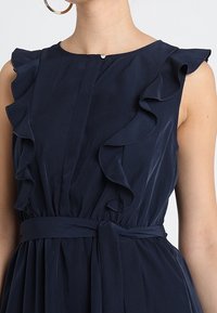 Mörkblå klänning med volangdetaljer vid axlarna, knappstängning vid halsen och ett matchande tygbälte i midjan. Slät textur.