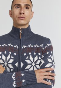 Dunkelblauer, gestrickter Reißverschluss-Pullover mit weißen und rostfarbenen Schneeflockenmuster. Rippkragen und -bündchen mit bronzenem Reißverschluss-Detail.