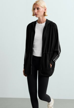 Vrouw met kort blond haar die een wit overhemd draagt, een zwarte zip-up hoodie met witte strepen op de mouwen, zwarte legging en witte sneakers.