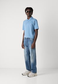 Pólo azul claro com mangas curtas e um pequeno logo quadrado, combinado com jeans de perna reta em lavagem clara e ténis cinzentos.