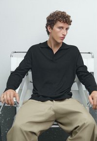 Svart långärmad polo med mjuk textur och V-halsdesign, tillsammans med beigefärgade vida byxor. Modell sitter i en stol av krom.