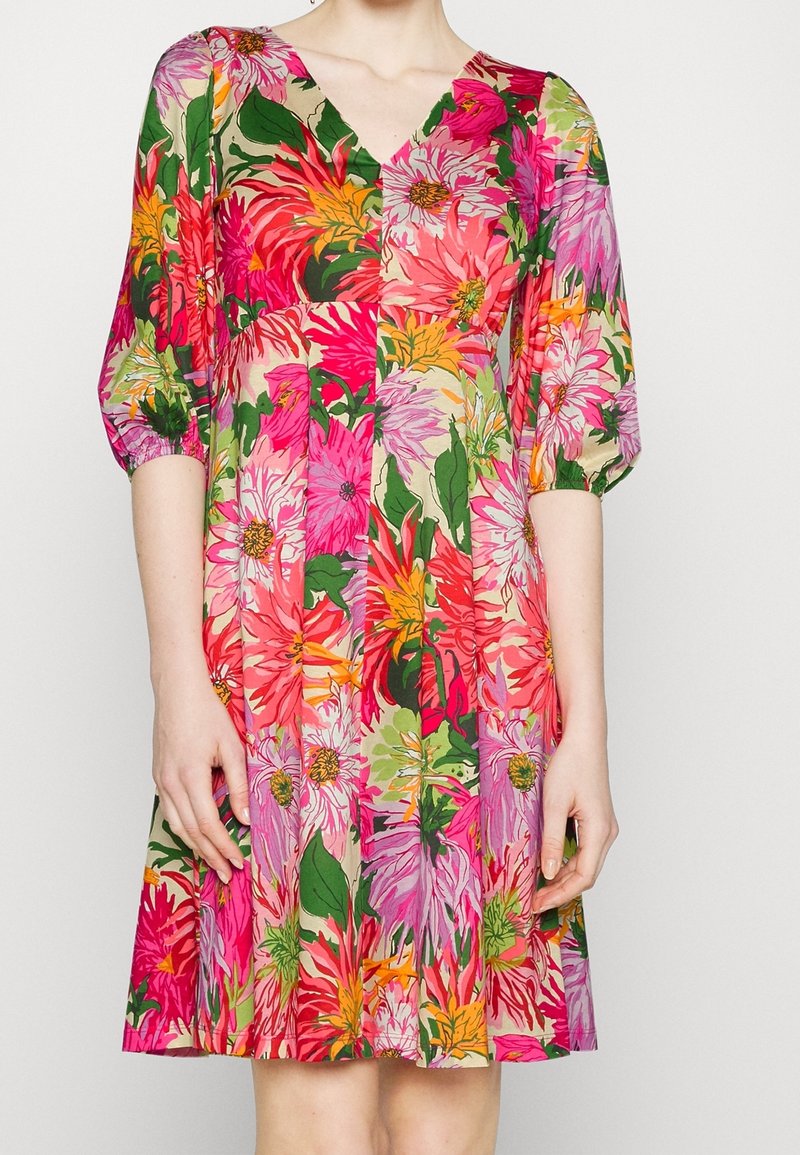 Femme portant une robe longueur genou avec un grand imprimé floral rose, rouge, jaune et vert et des manches bouffantes trois-quarts sur fond blanc.