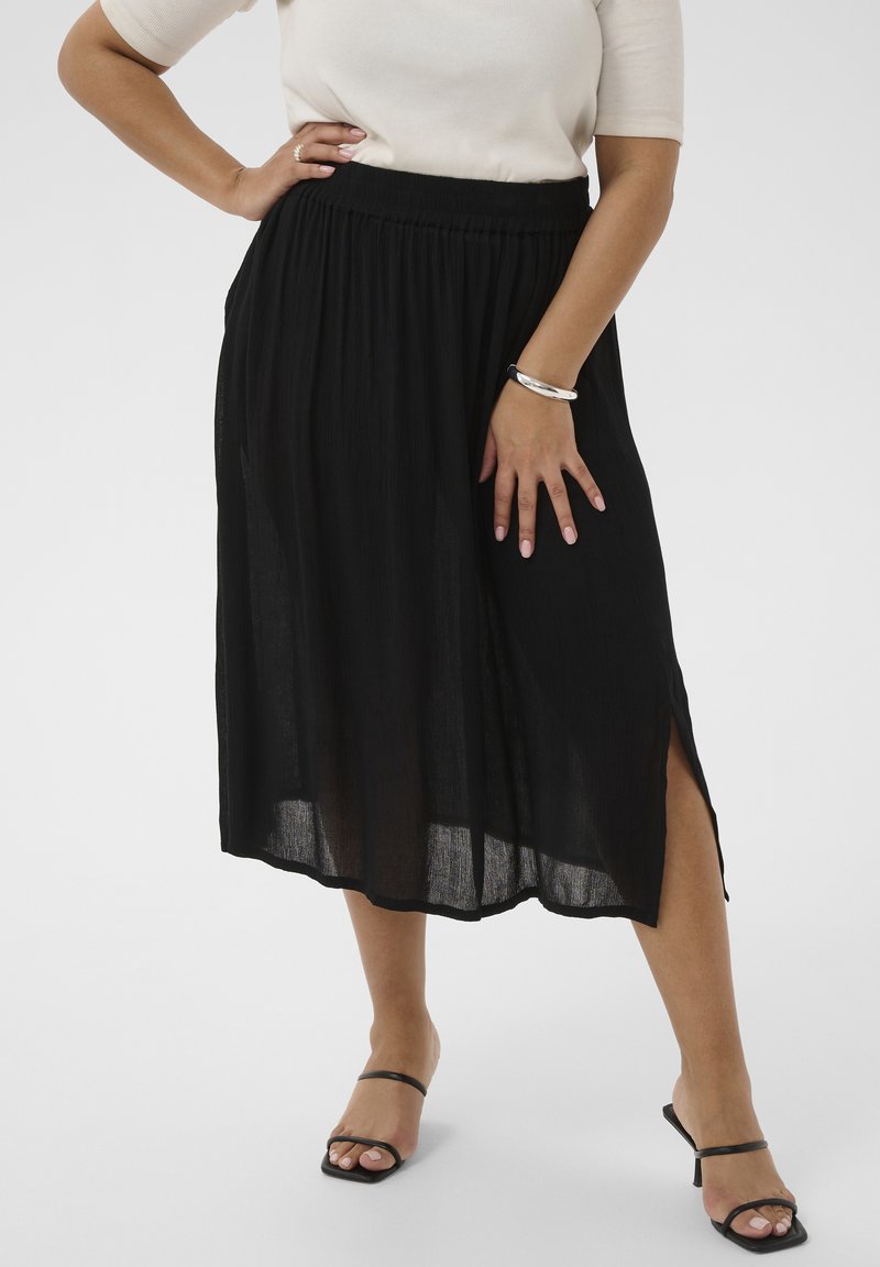 Femme debout, une main sur la hanche, portant une jupe midi noire avec une fente latérale, un haut blanc, un bracelet argenté et des talons ouverts noirs.