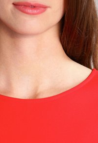 Czerwona bluzka z dekoltem w serek o gładkiej teksturze. Zbliżenie na dekolt i górną część klatki piersiowej, z miękkimi, naturalnymi odcieniami skóry i lekko błyszczącymi ustami.