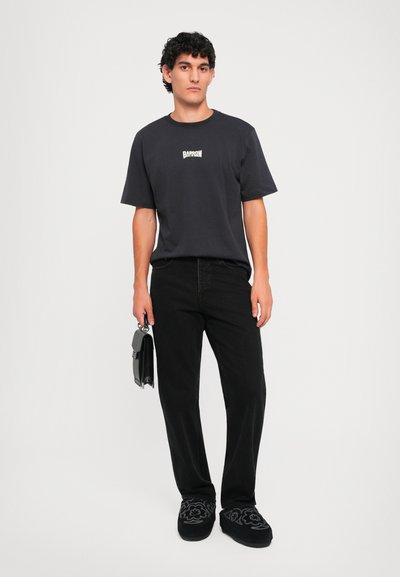 Hombre con camiseta negra con logo, pantalones negros y zapatos negros con estampado, sosteniendo un pequeño bolso negro, de pie contra un fondo blanco.