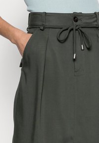 Gröna plisserade byxor med dragsko i midjan, med en framficka och ett slätt, texturerat tyg. Silverfärgade detaljer finns.