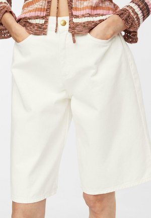 Mains dans les poches d’un short blanc taille haute, jambes larges, longueur genou, associé à un top court en maille multicolore à manches longues.
