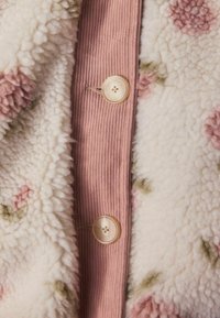 Fuzzy creme stof med blomster mønster, med en lyserød kordfløjlsstribe og to runde beige knapper med fire huller.