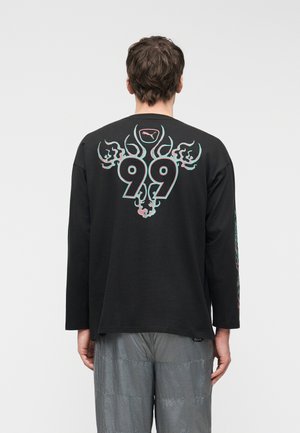 Man wearing black long-sleeve shirt with blue and pink flaming number "99" and Puma logo on the back, paired with gray pants.