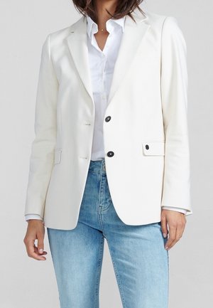 Žena v bílem na míru šitém saku s černými knoflíky přes bílou košili a světle modré džíny, stojící před jednobarevným pozadím.