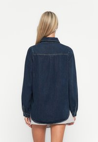 Dark blue denim shirt with long sleeves, button cuffs, and a classic collar. Features a rounded hem and subtle stitching details.