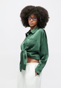 Πράσινο σατέν μπλουζάκι με δεμένη μπροστινή λεπτομέρεια, κουμπωτό σχέδιο και μία τσέπη στο στήθος. Συνδυάζεται με λευκά παντελόνια με πλίτσες.