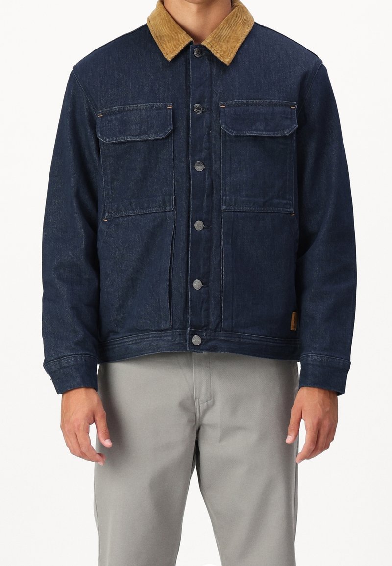 Tummansininen farkkutakki, jossa on vaaleanruskea samettikaulus, napit edessä ja kaksi rintataskua. Takissa on rento leikkaus ja teksturoitu kangas.