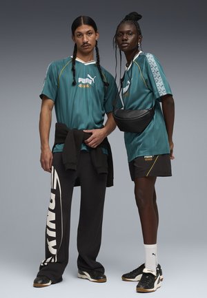 Dwoje dorosłych ubrane w morskie zielone sportowe ubrania Puma King, jedno z warkoczami i czarnymi spodniami, drugie z warkoczami, w szortach i z czarną torbą przewieszoną przez ramię.