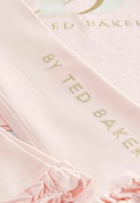 Gaiši rozā audums ar zelta izšūtu tekstu "TED BAKER" un "BY TED BAKER." Iezīmēts ar viļņotu apdari, gludu tekstūru un ikdienas dizainu.