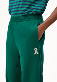 Pantalons de survêtement en coton vert avec une taille élastique, des poches latérales et un petit logo gris sur le bas de la jambe. Texture lisse et coupe décontractée.
