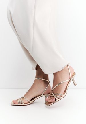 Lábak fényes, vékony pántos, magas sarkú szandálban, világos, bő szárú nadrággal, egyszínű fehér háttér előtt.