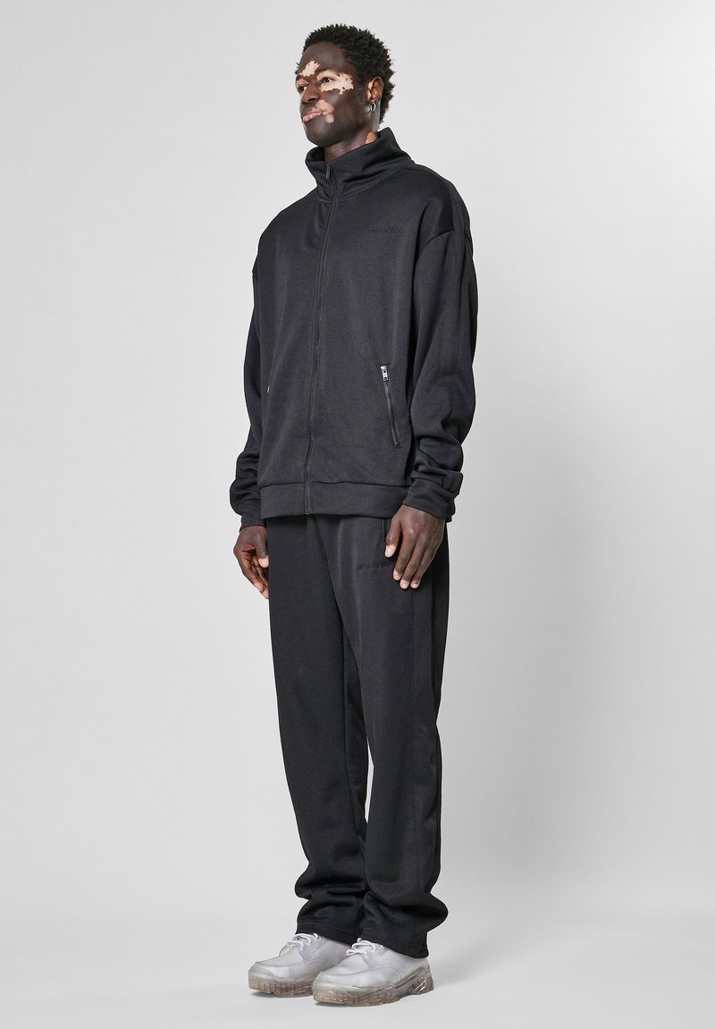 Melns zip-up džemperis ar augstu apkakli un sānu kabatām, savienots ar atbilstošiem melniem sporta biksēm. Modelis valkā gaišas kurpes.