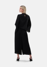 Frau in schwarzen, plissierten Hosen mit weitem Bein und durchsichtiger Bluse mit langen Ärmeln, steht mit in den Taschen steckenden Händen, seitlich stehend mit nach links gedrehtem Kopf.